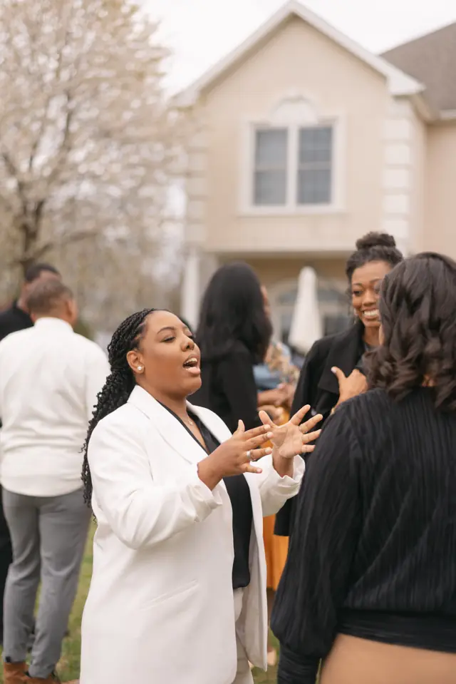 A Black woman in a white blazer speaks with her hands open while her sisters listen and smile.