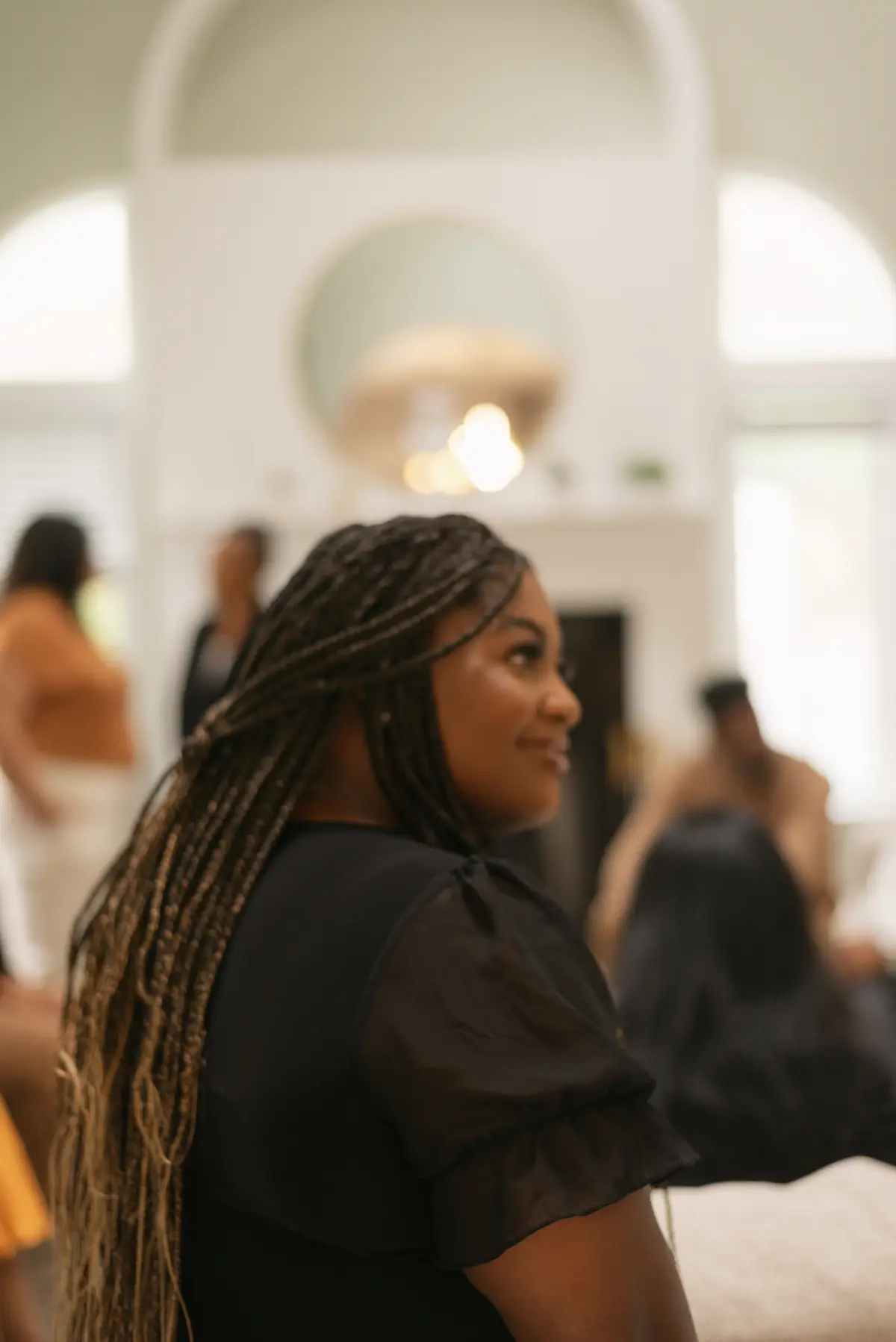 A Black woman with long braids stands in a sunlit room, looking up and smiling softly.